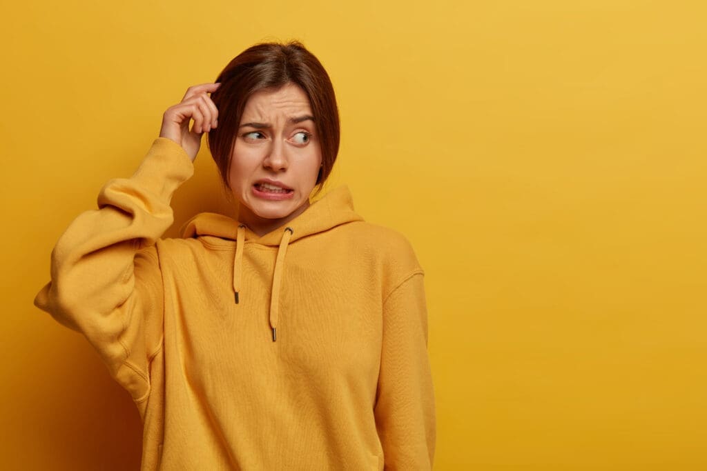 A woman, wearing a mustard-coloured hoodie, gently scratches her hair and makes an expression for- what is the best dry shampoo for oily hair.
