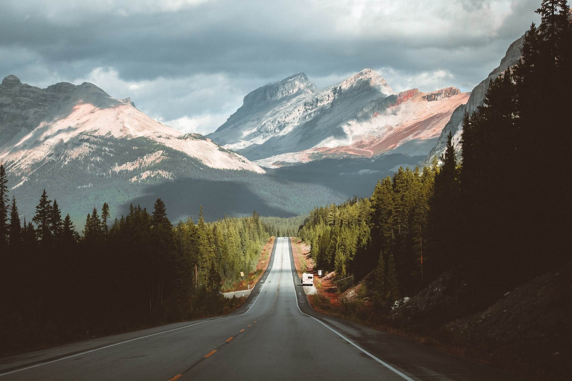 A grey concrete road between green trees and mountains during the daytime, Alberta, Canada.
