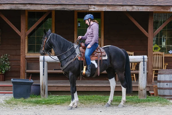 A young woman riding a black and white horse in front of a wooden house